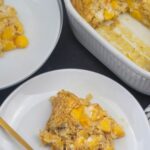 A serving of peaches and cream baked oatmeal on a white plate with a gold fork, next to a baking dish and another plate with more oatmeal.