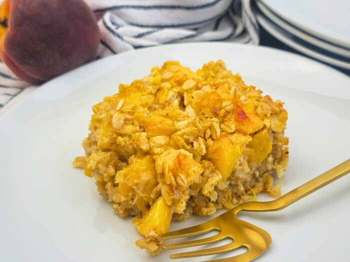 A square serving of baked oatmeal with peach pieces on a white plate, next to a gold fork, with a peach and stacked plates in the background.