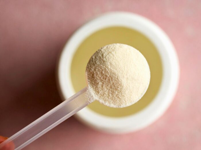 A close-up of a plastic spoon holding a scoop of fine, white powder above an open container on a pink surface.