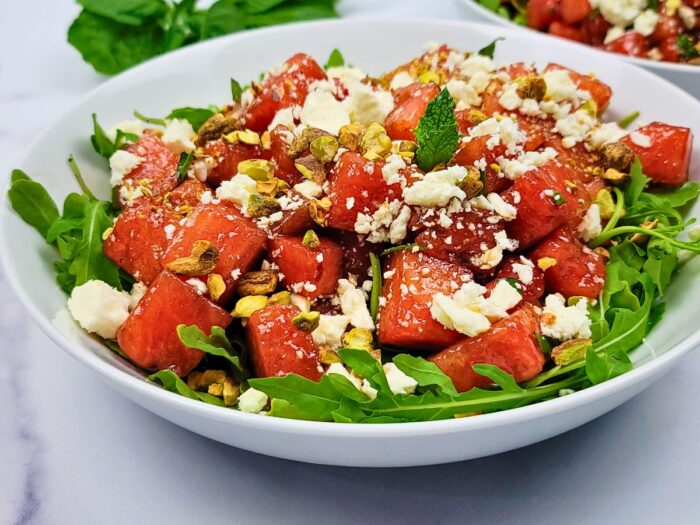 A bowl of salad with arugula, watermelon cubes, crumbled feta cheese, chopped pistachios, and mint leaves.