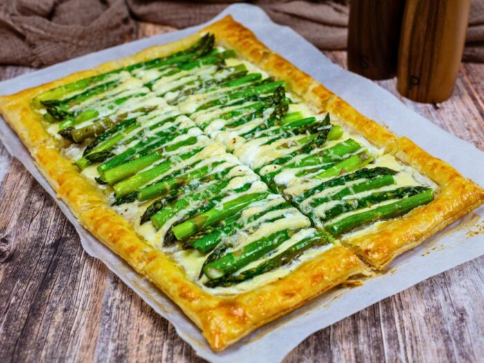 A rectangular puff pastry tart topped with asparagus spears and melted cheese, placed on parchment paper over a wooden surface.