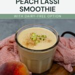 A glass of peach lassi smoothie topped with chopped pistachios and cinnamon sits on a wooden board next to a peach and a pink cloth. Text on image: Homemade Peach Lassi Smoothie.