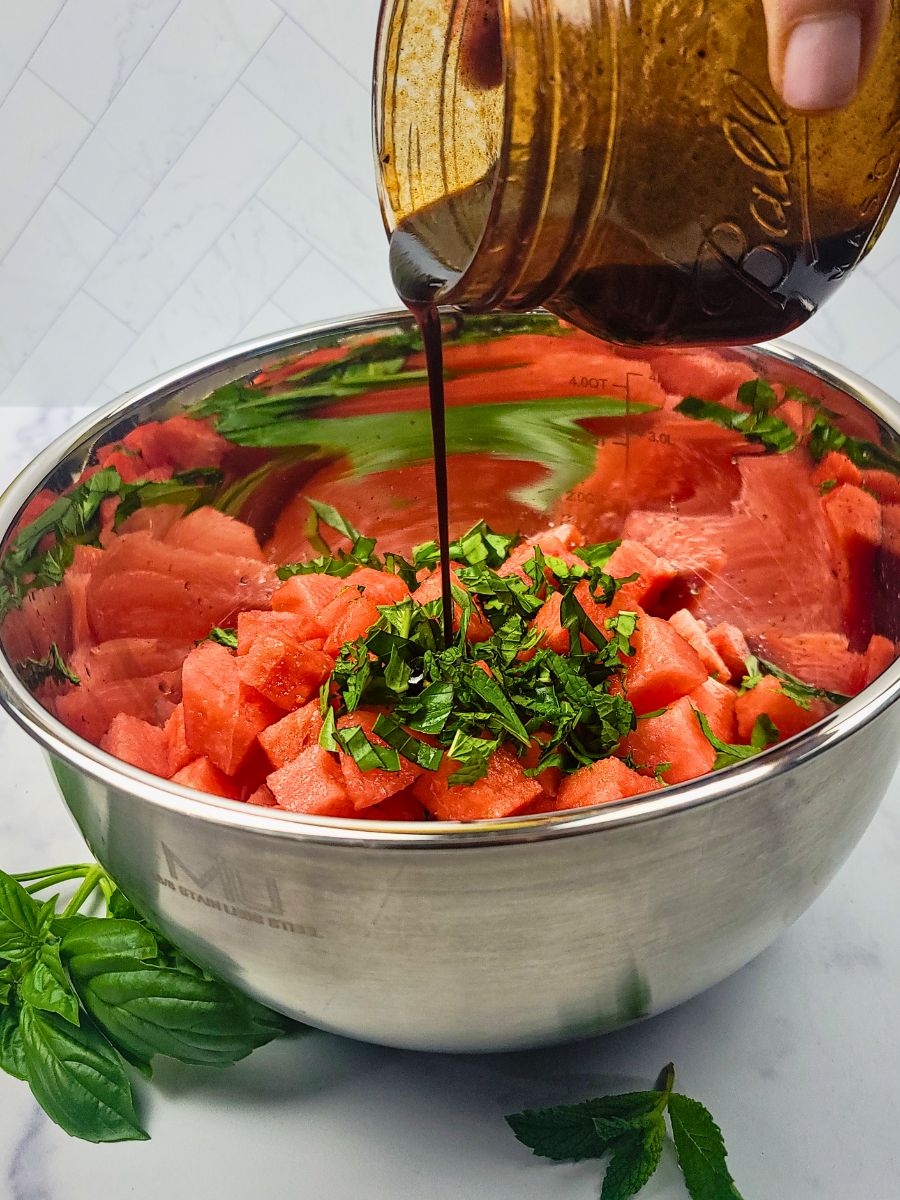 A hand pours dark dressing from a glass jar onto chopped watermelon and herbs in a large metal mixing bowl, with fresh herbs nearby.