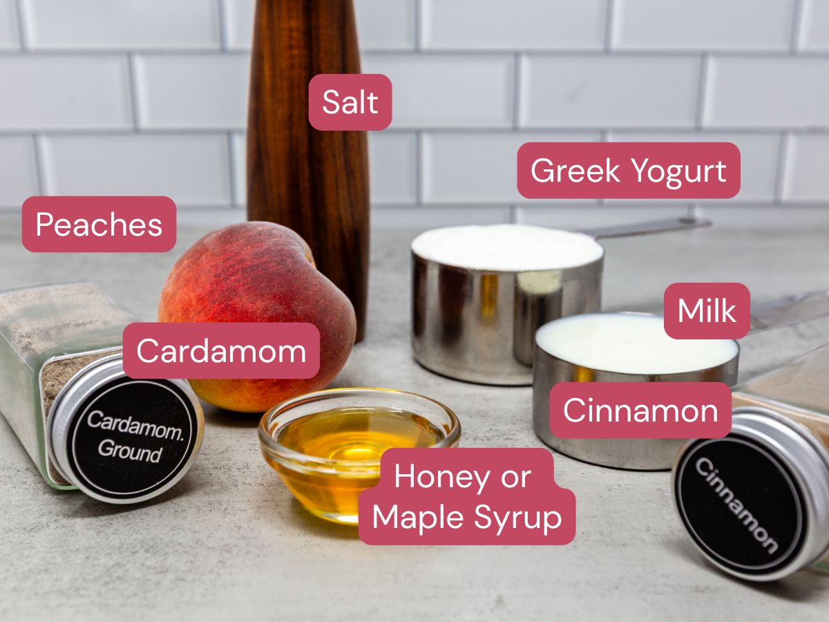 A peach, Greek yogurt, milk, honey or maple syrup, ground cardamom, ground cinnamon, and a salt shaker arranged on a countertop with labels.