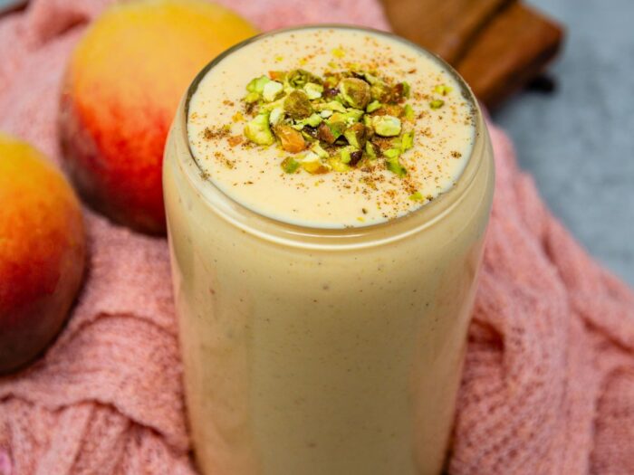 A glass of creamy peach smoothie topped with chopped pistachios and a sprinkle of cinnamon, with peaches and a pink cloth in the background.