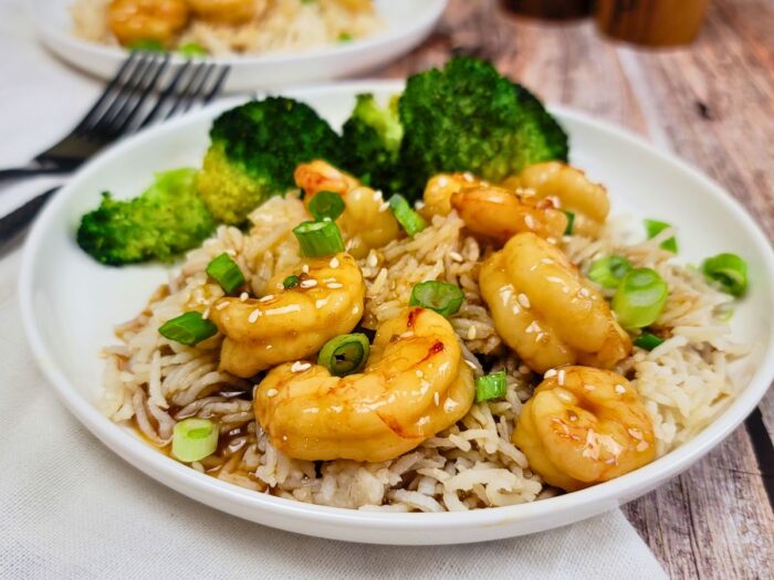 A plate of shrimp with sauce served over rice, garnished with green onions and sesame seeds, with steamed broccoli on the side.